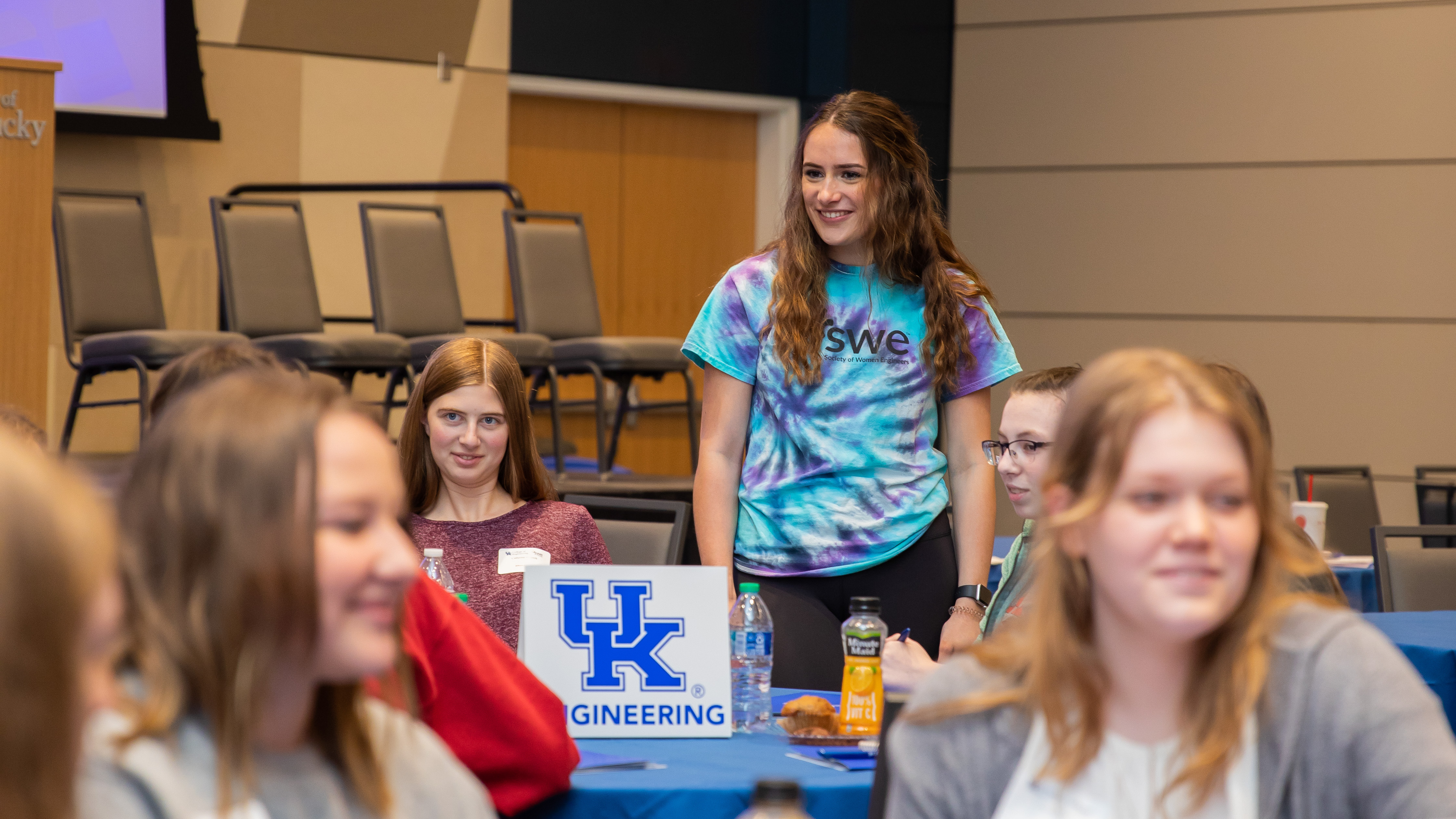 Student (female) with a tie-dyed SWE shirt standing at a table around a group of other female students.