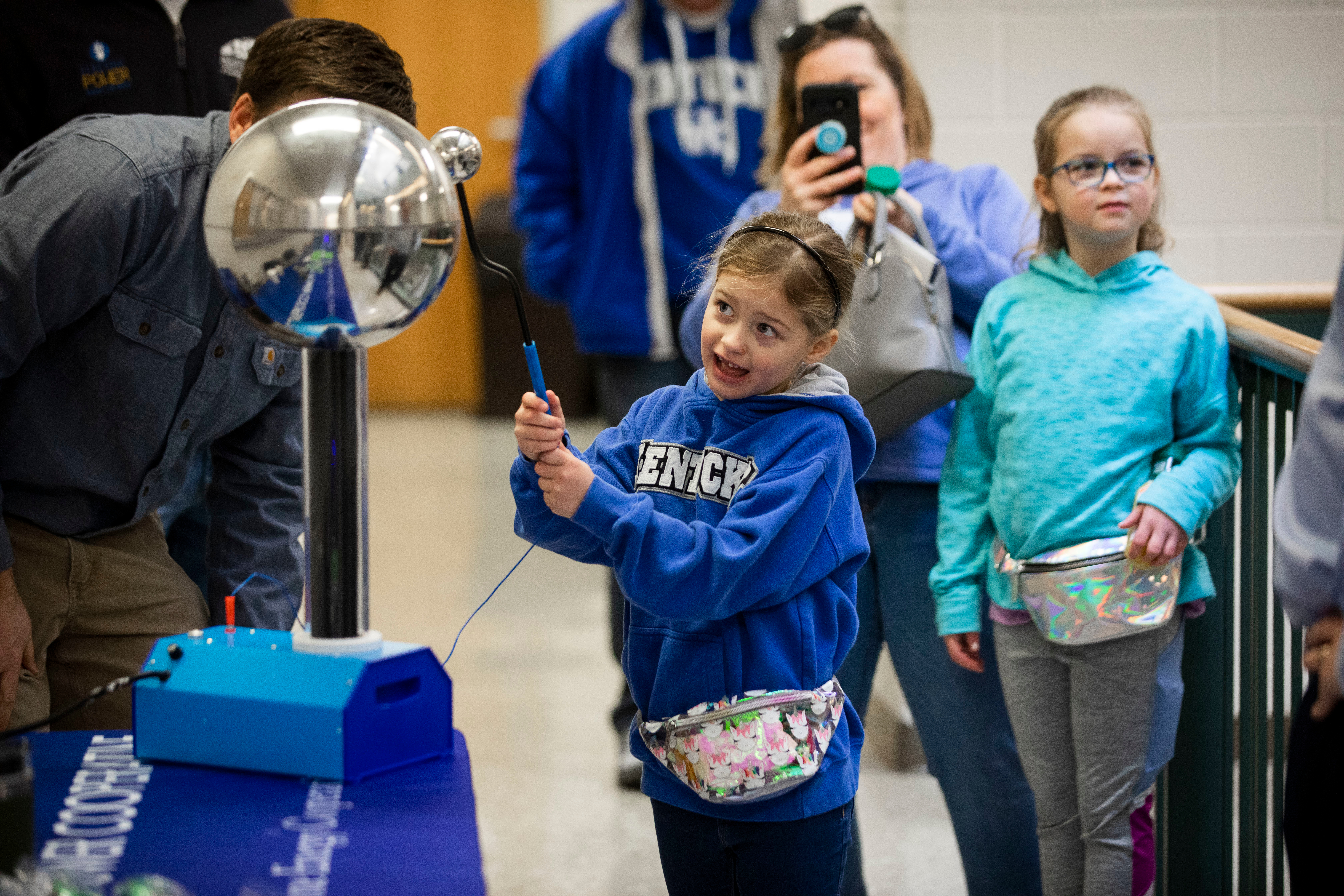 A young girl in a blue Kentucky hoodie interacts with a Van de Graaff generator, causing her hair to stand up, while people watch and take photos.