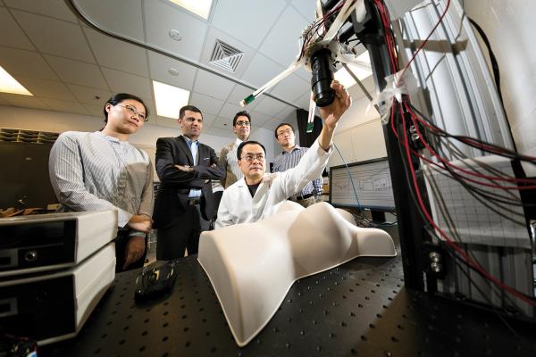 A group of five individuals in a laboratory setting observe a biomedical engineering experiment. At the center, a researcher in a lab coat adjusts a piece of equipment connected to wires and sensors. The setup includes a mannequin torso on a table with various technological components. The other individuals, dressed in business or casual attire, stand attentively in the background. Monitors and laboratory tools are visible in the workspace.