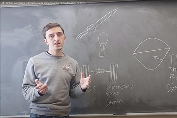 A person stands in front of a chalkboard giving a presentation. The chalkboard features drawings of a rocket, a parachute, the International Space Station, and a diagram labeled "TPS" for "Thermal Protection System." The presenter gestures with one hand, wearing a gray sweatshirt, while explaining the content.