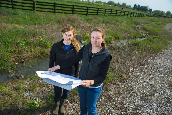 Two researchers outdoors holding blueprints, standing beside a small stream and surrounded by grass, with a fenced field in the background.