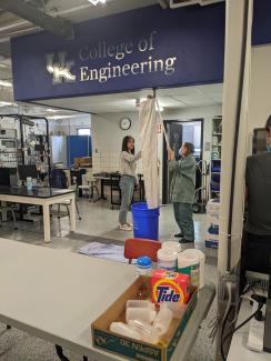 People holding white cloth up to a pipe in the ceiling into a blue recycling can under a College of Engineering sign.