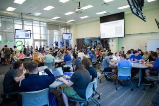 A classroom full of students sitting at tables and a projection screen that says "Engage with an engineer" at the front of the classroom.