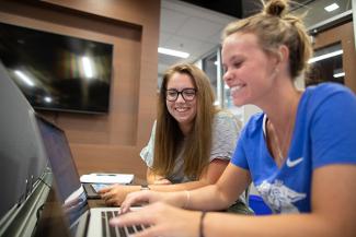 Students Caylee and Amanda smiling and laughing while working on laptops.