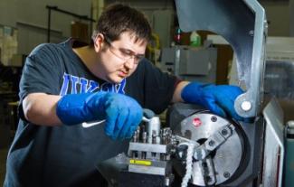 Student in large blue gloves working on a piece of manufacturing equipment.