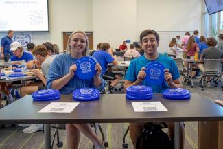 Two students holding up frisbees while sitting at a table at an event.