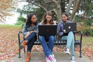 Three female students two with laptops working on a bench outside on a fall day.
