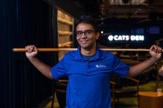 Student holding a pool queue, with a "Cats Den" sign behind him. 