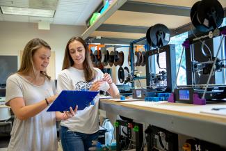 Two students working looking at a clipboard while working on 3D printers.