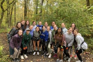 Group of women in the woods, wearing hiking and climbing gear with fall colored leaves around them.