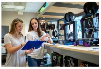 Students in MakerLab space looking at a clipboard surrounded by 3D printers.