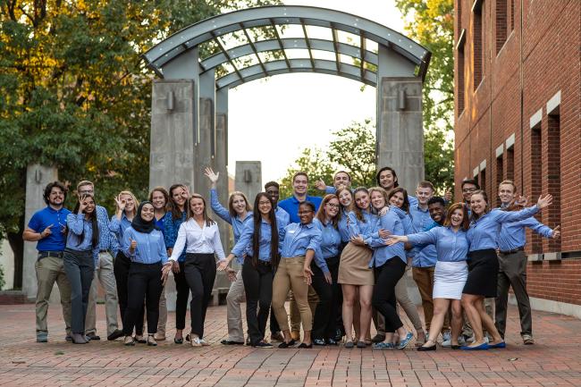 Group of Engineering Ambassadors wearing matching button down shirts, standing underneath the Engineering Arches with in the Engineering complex with some students having their arms spread wide open.