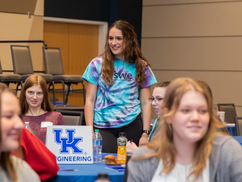 Student (female) with a tie-dyed SWE shirt standing at a table around a group of other female students.