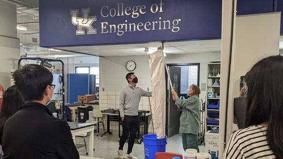 People holding white cloth up to a pipe in the ceiling into a blue recycling can under a College of Engineering sign.