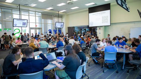 A classroom full of students sitting at tables and a projection screen that says "Engage with an engineer" at the front of the classroom.