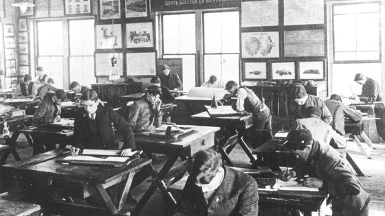 Black and white image of all male students sitting at desks with a sign that says "School of Mechanical Engineering, State College of Kentucky" on the wall.