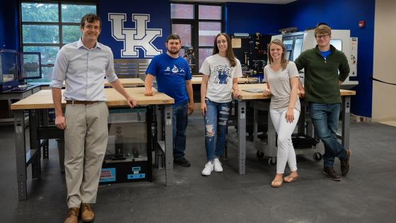 Doug Klein and 4 students posing in the innovation center with 3D printers in the background.