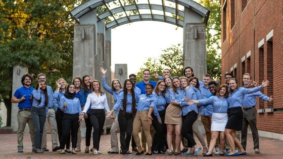 Group of Engineering Ambassadors wearing matching button down shirts, standing underneath the Engineering Arches with in the Engineering complex with some students having their arms spread wide open.