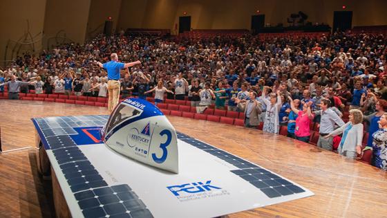 Solar car in the foreground of the image, on a stage with Tony Jackson in the background animatedly engaging an auditorium full of people. 