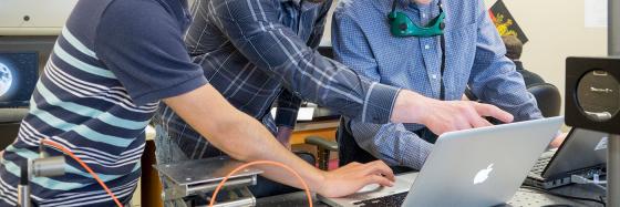 3 students in a lab pointing at a MacBook.