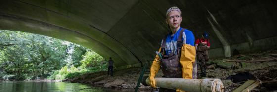 Students wearing waders holding a PVC and metal contraption in a river under a bridge.