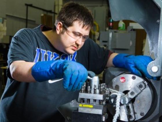 Student in large blue gloves working on a piece of manufacturing equipment.