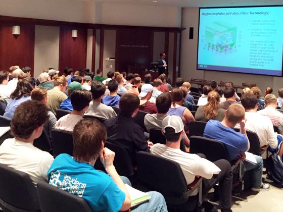 Students in a large auditorium, viewing a presentation given on a projector screen at the front of the room.