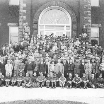 Black and white picture of alumni from around the 1940's, with mostly all men standing on the steps of a building, wearing suits or military uniforms and one woman in the second row.